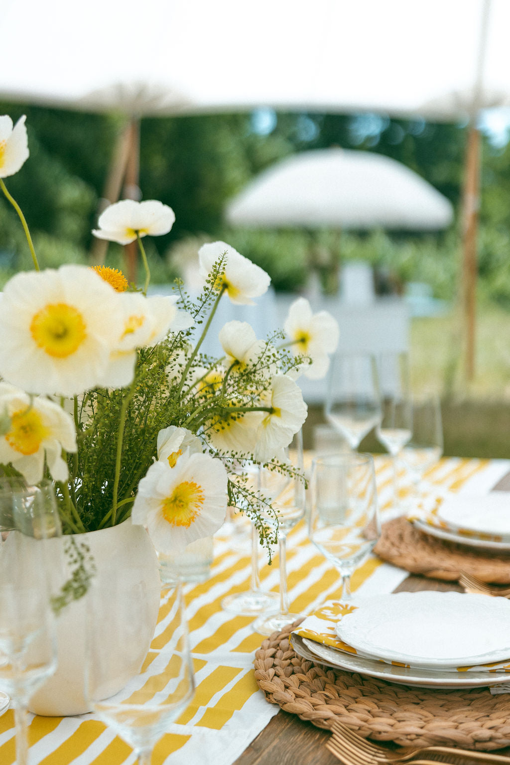 Outdoor table setting with white flowers, yellow and white striped Erin Flett tablecloth, and white plates.