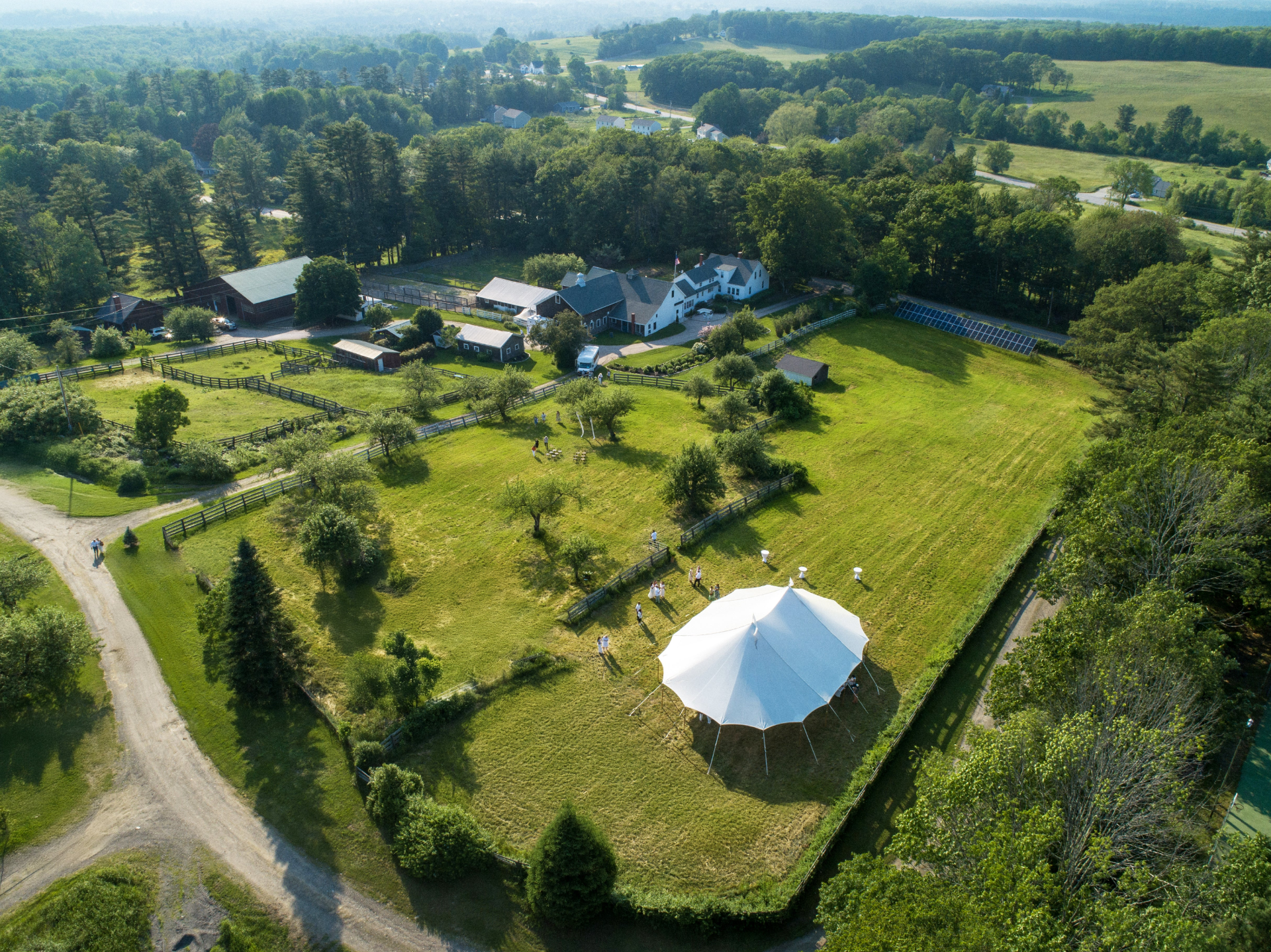 Aerial view of a white tent on a grassy field with trees and buildings in the background