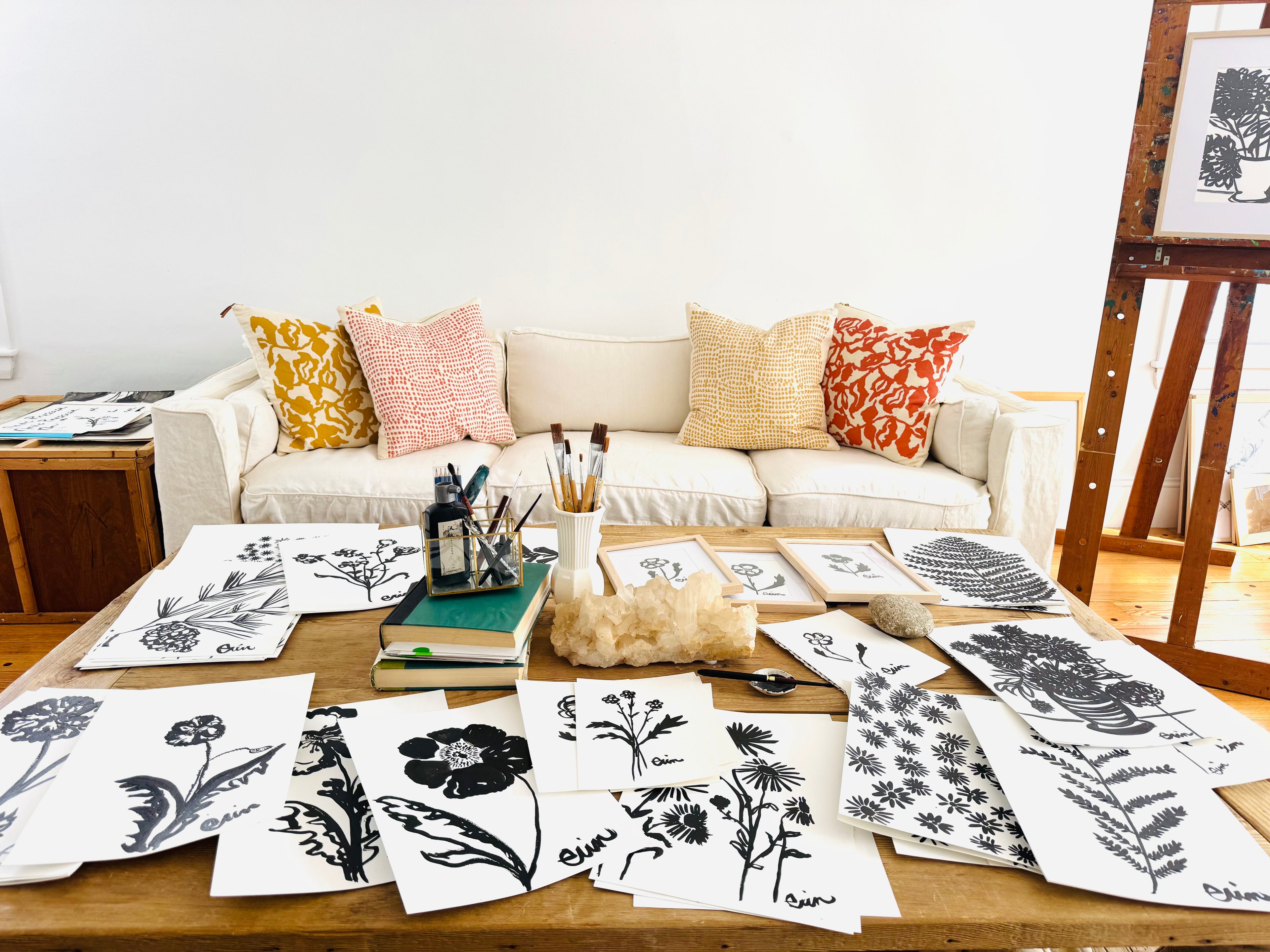 Table with various black and white Erin Flett prints of floral and abstract designs, a couch with patterned cushions in the background.