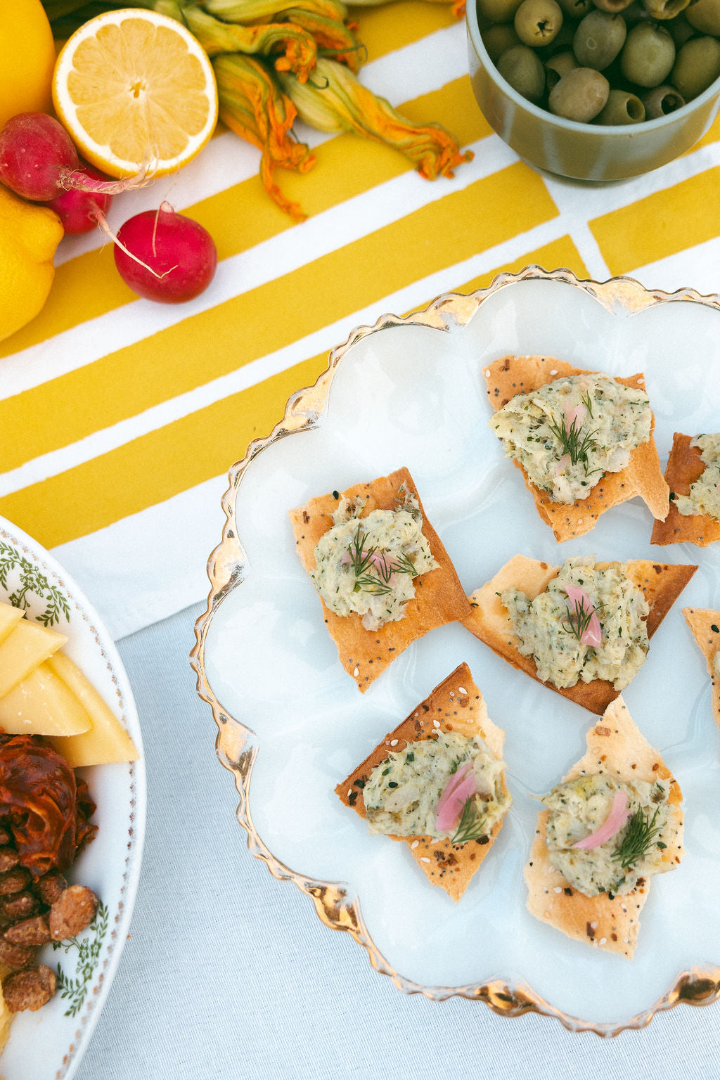 Plated appetizers with crackers topped by a spread, surrounded by fruits and vegetables on a striped tablecloth.