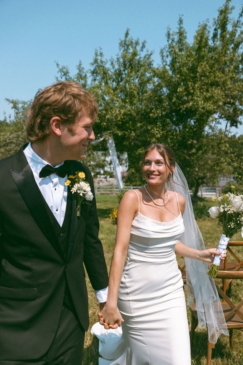 Man in a tuxedo and woman in a white dress holding hands outdoors with trees and a clear sky in the background.